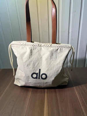 Beige shoppingväska från alo - Snygg shoppingväska i beige tyg med bruna axelband i konstläder och dragsko upptill. Stor svart alo-logga framtill. Perfekt för att bära allt du behöver till skolan, gymmet eller stan. Rymmer mycket och har en clean, modern look.