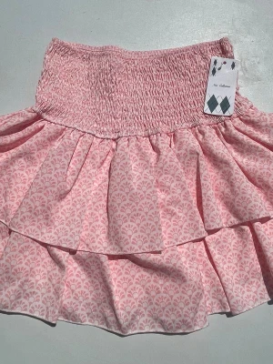 Rosa volangkjol med mönster - Söt kortkjol i ljusrosa med volanger och ett diskret blommigt mönster. Kjolen har smock i midjan för en bekväm passform och är gjord i ett lätt bomullstyg som passar perfekt till varmare dagar. Köpt i Frankrike. Har inbyggda shorts.