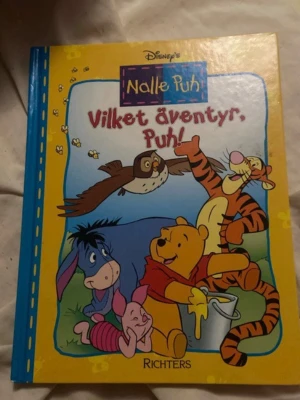 Nalle Puh: Vilket äventyr, Puh! - En färgglad barnbok med hårda pärmar från Disney om Nalle Puh och hans vänner. Omslaget är gult och blått med illustrationer av Puh, Tiger, Ior, Nasse och Uggla. Boken är utgiven av Richters och handlar om ett spännande äventyr i Sjumilaskogen.