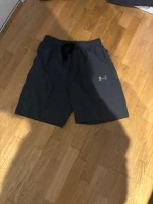 Gråa träningsshorts från Under Armour - Svarta träningsshorts från Under Armour med elastisk midja och snörning. Färgglad logga framtill och detaljer i flera färger baktill. Perfekta för sport eller gym och tillverkade i lätt syntetmaterial.