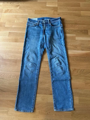 Blå raka jeans i klassisk modell - Säljer ett par blå jeans med rak passform och normal midja. Jeansen har fem fickor, bälteshällor och dragkedjegylf. Materialet är klassisk denim i bomull och färgen är mellanblå med lätt slitning. Perfekta till en avslappnad stil.