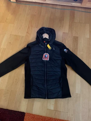 Svart jacka från Parajumpers helt ny - Snygg svart pufferjacka från Parajumpers med huva och dragkedja framtill. Jackan har quiltade paneler, två sidofickor och en gul detalj vid halsen. Perfekt för kalla dagar och har en sportig look.
