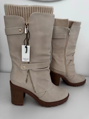 Beige höga stövlar med klack - Snygga beige stövlar med grov klack och dekorativa guldfärgade dragkedjor. Stövlarna har ribbstickad kant upptill och en remdetalj över foten. Tillverkade i ett matt material som liknar mocka och har en robust sula i brunt. Perfekta för höst och vinter.
