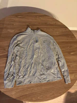 Ljusgrå halvzip tröja Ralph Lauren - Ljusgrå långärmad tröja med halv dragkedja och diskret broderad logga på bröstet. Tröjan har ribbade muddar vid ärmslut och nederkant, perfekt för en avslappnad och stilren look.