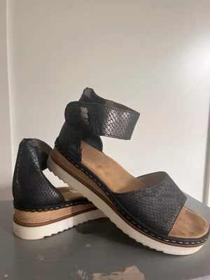 Svarta sandaler med ormmönster Rieker - Snygga svarta sandaler från Rieker med trendigt ormmönster och breda remmar runt vristen. Sandalerna har en vit, räfflad platåsula och en bekväm innersula i beige mockaimitation. Perfekta för dig som vill ha både stil och komfort.