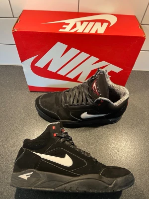 Nike svarta high top sneakers - Svarta Nike sneakers med högt skaft och vit swoosh på sidan. Skorna har svarta snören, detaljer i rött och grått samt mönstrat innerfoder. Ovandelen är i mockaliknande material och sulan är svart med vit logga på hälen. Perfekta för dig som gillar retrostil.