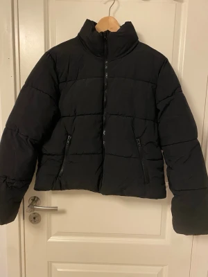 Svart pufferjacka med hög krage - Snygg svart pufferjacka med hög krage och dragkedja framtill. Jackan har två praktiska fickor med dragkedja och quiltad design som ger en cool look. Perfekt för kalla vinterdagar och riktigt skön att bära. Materialet är syntet och jackan har en boxig passform.