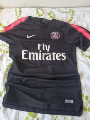 Svart PSG matchtröja från Nike - Säljer en svart PSG matchtröja från Nike med vita loggor och texten Fly Emirates på bröstet. Tröjan har rosa detaljer på axlarna och är tillverkad i ett lätt och ventilerande material, perfekt för fotboll eller träning.