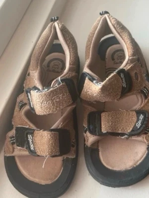 Beige sandaler med kardborre(barn)  - Säljer ett par beige sandaler med svarta detaljer och kardborreband. Sandalerna har öppen tå och häl, samt texten 'Sport' på banden. Sulan är svart och materialet ser ut att vara mockaimitation och textil.