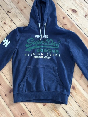 Mörkblå Superdry hoodie med tryck - Mörkblå hoodie från Superdry med stor grön och vit logga på bröstet och japansk text. Har huva med vita snören och en stor magficka. På vänster ärm står det 'JPN' i vitt. Perfekt för en avslappnad streetwear-look.