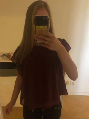 Vinröd blus med volangärmar - Säljer en vinröd blus i luftig och lätt material med volangdetaljer på ärmarna och spets vid bröstet. Blusen har rund halsringning och korta ärmar.💕