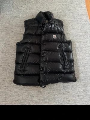 Svart dunväst från Moncler - Snygg svart dunväst från Moncler med hög krage och två dragkedjefickor framtill. Västen har ett glansigt material och klassisk Moncler-logga på bröstet. Insidan har ett serietidningsmönster och etikett med QR-kod. Västen är köpt från Care of Carl för nypris