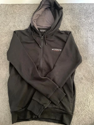 Svart zip hoodie från McKenzie - Snygg svart hoodie från McKenzie med dragkedja och huva. Hoodien har ribbade muddar, fickor på sidorna och logga tryckt på bröstet. Insidan av huvan är grå. Perfekt för en chill och sportig look.