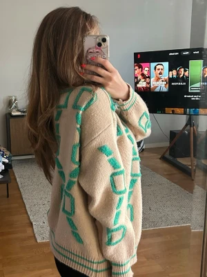 Beige kofta med turkost mönster - Säljer en beige kofta med turkosa geometriska mönster och ribbade muddar. Koftan har knappar framtill och en avslappnad passform. Perfekt för dig som gillar färgglada detaljer och unik design.
