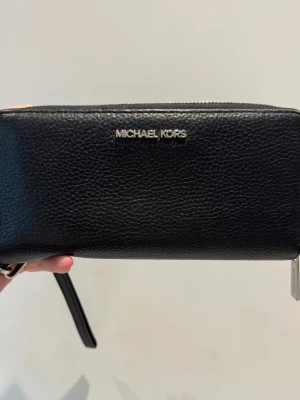 Super fin! Svart plånbok från Michael Kors - Snygg svart plånbok i äkta läder från Michael Kors med silverfärgad logga framtill. Plånboken har dragkedja runtom, många kortfack, fönster för ID och ett handledsband. Perfekt för att hålla ordning på kort och kontanter. Knappt använd i super fint skick! Det enda man kan anmärka på är att dragkedja flärpen har några ytliga repor, syns om man granskar noga i rätt ljus. Nypris 1800:-