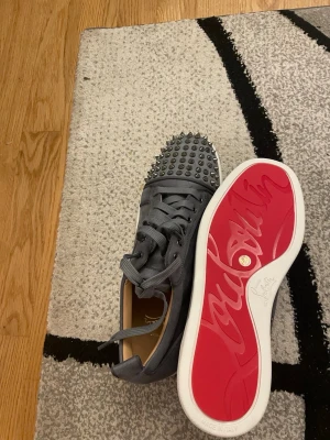 Gråa sneakers med nitar och röd sula - Snygga gråa sneakers med matta nitar på tån och klassisk röd sula. Skorna har snörning och är tillverkade i mockaliknande material med vita kanter på sulan. Perfekta för dig som vill sticka ut med en edgy och lyxig stil.