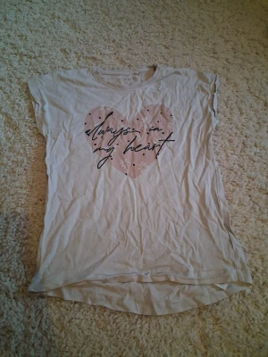 Vit t-shirt med hjärta och text - Söt vit t-shirt med ett stort rosa hjärta och texten 'always in my heart' framtill. T-shirten har korta ärmar och rund halsringning. Perfekt för dig som gillar gulliga tryck och enkel stil.