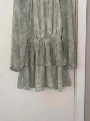 Ljusgrön blommig kortklänning - Söt ljusgrön kortklänning med vitt blommönster och volangdetaljer. Klänningen har långärmade ärmar och resor i midjan för en bekväm passform. Perfekt för vår och sommar med sitt luftiga material. Har använt den en till två gånger men man ser knappt att den är använd.