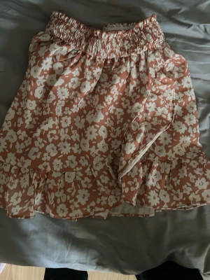 Blommig kortkjol med volang - Söt kortkjol med volangkant och omlottkänsla i ljusrosa och vitt blommönster. Kjolen har bred resår i midjan för skön passform och är perfekt för vår och sommar. Tillverkad i ett lätt och luftigt material.