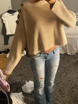 Beige ribbstickad croppad tröja - Trendig beige ribbstickad tröja med croppad passform och stora dekorativa knappar längs ena axeln. Tröjan har långa ärmar och en bred, rak halsringning. Perfekt för dig som gillar en avslappnad men snygg stil.