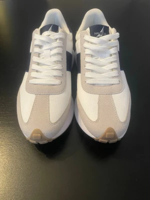 Vita och beige Nautica sneakers - Snygga sneakers från Nautica i vitt och beige med mörkblå detaljer och logga på hälen. Ovandelen är i textil och mockaliknande material, med vita snören och en klassisk låg siluett. Perfekta för en stilren och sportig look.