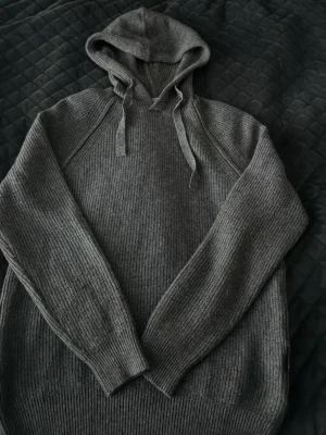 Grå stickad hoodie från Replay - Mysig grå stickad hoodie från Replay med ribbstickad struktur och dragsko i huvan. Tröjan har långa ärmar och en avslappnad passform, perfekt för en chill och stilren look. Materialet känns mjukt och exklusivt.
