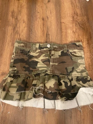 Camouflagekjol med volang från Gina Tricot - Säljer en kort camouflagekjol från Gina Tricot i storlek 164. Kjolen har volang nertill, råa kanter och klassiska jeansdetaljer med fickor fram och bak. Perfekt för dig som vill ha en edgy och trendig look.