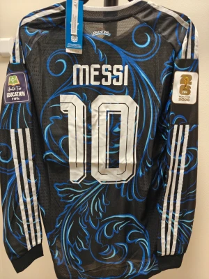 Argentina Messi långärmad  spelar version VM borta - Snygg långärmad Argentina matchtröja med Messi och nummer 10 på ryggen. Tröjan är svart med blått och vitt mönster, har vita ränder på ärmarna och officiella FIFA och AFA-emblem på bröstet. Tillverkad i ett lätt och ventilerande material.