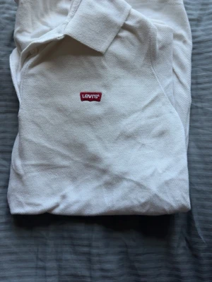 Vit pikétröja från Levi's - Klassisk vit pikétröja från Levi's med liten röd logotyp på bröstet. Tröjan har krage, korta ärmar och knäppning vid halsen. Tillverkad i mjuk bomull som känns skön mot huden. Perfekt till jeans eller shorts.