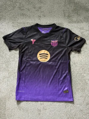 Svart-lila FC Barcelona matchtröja - Snygg FC Barcelona matchtröja i svart och lila med guldiga detaljer. Tröjan har korta ärmar, klubbmärke och sponsorlogga framtill samt Lamine Yamal och nummer 10 på ryggen. Tillverkad i ett lätt och ventilerande funktionsmaterial.