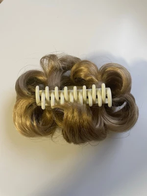 Blond hårklämma med syntetiskt hår - Säljer en hårklämma med fastsatt lockigt syntetiskt hår i en naturlig blond nyans. Perfekt för att snabbt fixa en voluminös frisyr. Klämman är i vit plast och håret är glansigt och mjukt. Aldrig använd, endast testad. 