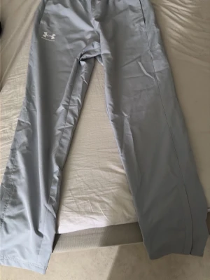 Ljusgrå träningsbyxor Under Armour - Ljusgrå träningsbyxor från Under Armour med rak passform och elastisk midja. Byxorna har en diskret logga på vänster lår och är tillverkade i ett lätt och smidigt syntetmaterial som passar perfekt för sport och aktivitet.