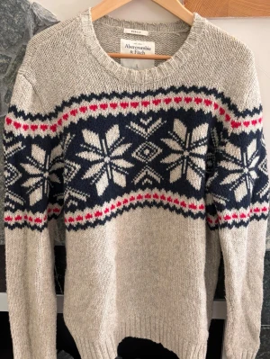 Beige stickad Abercrombie & Fitch tröja med mönster - Stickad tröja från Abercrombie & Fitch i beige med mörkblått och vitt snöflingemönster samt röda detaljer. Rund halsringning och ribbade muddar vid ärmslut och nederkant. Perfekt för kalla dagar och riktigt mysig stil.