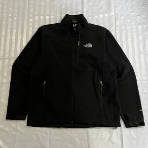 Svart vindjacka från The North Face - Snygg svart vindjacka från The North Face med hög krage och dragkedja framtill. Jackan har tre fickor med dragkedja och diskret logga på bröstet. Perfekt för blåsiga dagar och enkel att matcha med olika outfits.