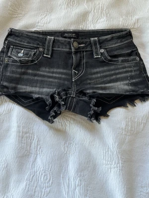 Svarta jeansshorts True Religion - Säljer ett par svarta jeansshorts från True Religion med slitna kanter och markerade vita sömmar. Shortsen har fem fickor, knappgylf och coola detaljer på bakfickorna. Perfekta för sommaren och ger en edgy vibe till din outfit.