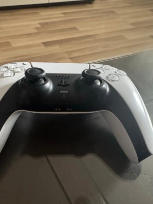 Vit och svart PS5 DualSense handkontroll - Säljer en snygg PS5 DualSense handkontroll i vit och svart. Den har klassisk PlayStation-design med touchpad, två analoga spakar, styrkors och de ikoniska symbolknapparna. Tillverkad i plast med ergonomisk form för skönt grepp under långa spelsessioner.