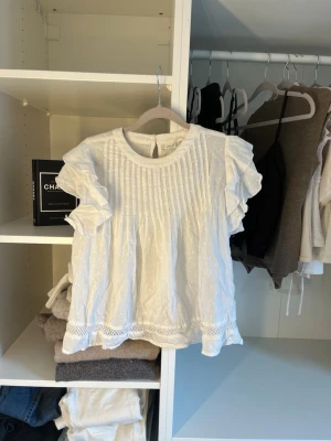 Vit blus med volang  - Söt vit blus med korta volangärmar, rund halsringning och fina spetsdetaljer längst ner. Blusen har diskreta prickar i tyget och knäppning bak i nacken. ganska stor i storleken💖