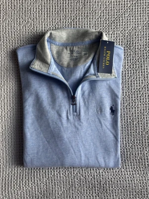 Ralph Lauren - Polo Ralph Lauren Quarter-zip i en fin pastelblå färg 🔵. Tröjans tyg är jättelen och skön att bära. Passar perfekt för att bygga en lagom seriös men också avslappnad look. Utmärkt som övertröja med skjorta eller piké under. Aldrig använd ✅ Priset är inte hugget i sten, så hör av dig gärna🤩