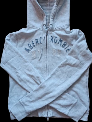 Ljusgrå hoodie med dragkedja - Ljusgrå hoodie från Abercrombie & Fitch med dragkedja framtill och broderad text i blått och vitt över bröstet. Tröjan har huva och dragskedja. Skriv vid frågor.