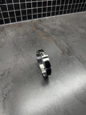 Svart och silverfärgat metallarmband - Stilrent armband i metall med svart emalj och silverfärgade detaljer. Armbandet har en modern design med ett H-format spänne framtill. Perfekt accessoar för att lyfta din outfit med en elegant touch.
