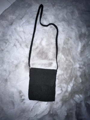 Svart virkad axelväska - En svart axelväska i virkat material med enkel, fyrkantig form och lång axelrem. Perfekt för dig som gillar en stilren och minimalistisk look. Väskan har ett lock framtill och är lätt att matcha med olika outfits.