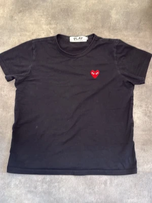 Svart t-shirt Comme des Garçons Play - Svart t-shirt från Comme des Garçons Play med det ikoniska röda hjärtat broderat på bröstet. Klassisk rund hals och korta ärmar. Tillverkad i mjuk bomull för en skön känsla och enkel stil.