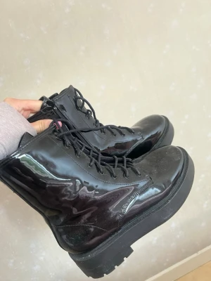Svarta lackade boots från Nelly storlek 37 - Snygga svarta boots i glansigt lackmaterial med kraftig platåsula och snörning framtill. Dragkedja på insidan för enkel på- och avtagning. Perfekta för att ge outfiten edge och attityd.