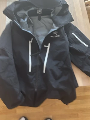 Svart vindjacka från Arc'teryx - Snygg svart vindjacka från Arc'teryx med vita dragkedjor och logga på bröstet. Jackan har huva, två bröstfickor med dragkedja och en ficka på ärmen. Tillverkad i slitstarkt material, perfekt för blåsiga dagar.