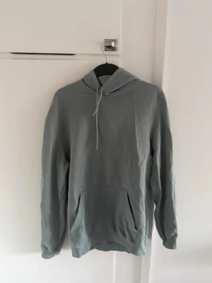 Gråblå hoodie - Säljer en gråblå hoodie med huva och dragsnören. Hoodien har en stor magficka framtill och ribbade muddar vid ärmslut och nederkant. Perfekt för en chill och avslappnad stil. Materialet känns mjukt och bekvämt, troligen bomull eller bomullsblandning.