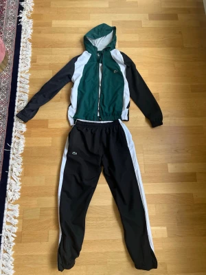 Grön och svart träningsdress Lacoste - Snygg träningsdress från Lacoste i grönt, svart och vitt. Setet består av en huvad jacka med dragkedja och matchande byxor med resår i midjan. Båda delarna har vita detaljer längs sidorna och den klassiska Lacoste-loggan på bröstet och benet. Perfekt för sport eller chill.