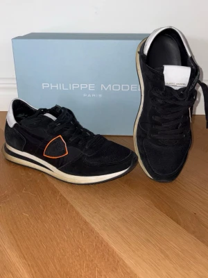 Svarta sneakers Philippe Model - Snygga svarta sneakers från Philippe Model med vita och orange detaljer. Skorna har en klassisk låg siluett, svart mocka och mesh samt vit sula med svarta och beige inslag. Perfekta för en trendig streetwear-look.