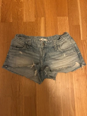 Ljusblå slitna jeansshorts - Säljer ett par ljusblå jeansshorts med slitna detaljer och råa kanter. Shortsen har fem fickor, bälteshällor och knappgylf. På bakfickorna finns snygga broderade detaljer. Perfekta för sommaren! Har ett litet slätt längst ner undertill men inget som påverkar.