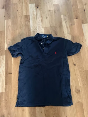 Mörkblå pikétröja från Polo Ralph Lauren - Klassisk mörkblå pikétröja från Polo Ralph Lauren med korta ärmar och krage. Tröjan har knappar framtill och en röd broderad logga på bröstet. Perfekt för en avslappnad och stilren look. Från ralph lauren