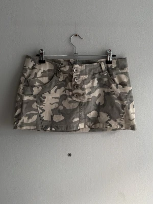 Camo minikjol med knappar - Säljer en kort camouflagemönstrad minikjol med fyra knappar framtill och stora fickor både fram och bak. Kjolen har bälteshällor och är gjord i ett slitstarkt bomullstyg. Perfekt för dig som gillar streetwear och utility-vibe. Litet hål där bak men går att laga enkelt 